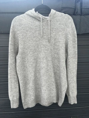 Ljusgrå Kashmir tröja - Mysig ljusgrå kashmir hoodie med huva och dragsnören. Tröjan har ribbade muddar vid ärmslut och nederkant, och är tillverkad i ett mjukt material som känns skönt mot huden. Perfekt för en avslappnad och stilren look. Storlek S passar mig som är 178 och väger 62 finns ett liten hål i tröjan som syns på bilden men märks inte av när man har på sig den