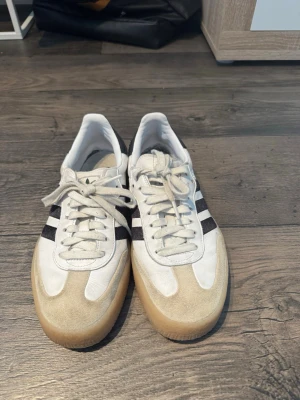 Adidas Samba vita sneakers - Klassiska Adidas Samba sneakers i vitt läder med svarta ränder på sidorna och beige mockadetaljer framtill. Skorna har en ikonisk gummisula och lågt skaft. Perfekta för en stilren och sportig look.