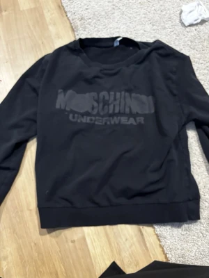 Svart loungewear-tröja Moschino - Svart långärmad loungewear-tröja från Moschino med tryckt logga 'Moschino Underwear' på bröstet. Tröjan har rund halsringning och är tillverkad i mjukt material som passar perfekt för chill dagar hemma.