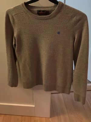 Beige stickad tröja från Morris - Snygg beige stickad tröja från Morris med rund halsringning och diskret blå logga broderad på bröstet. Tröjan har långa ärmar och ribbade muddar vid ärmslut och nederkant. Perfekt för lager-på-lager och enkel att matcha.