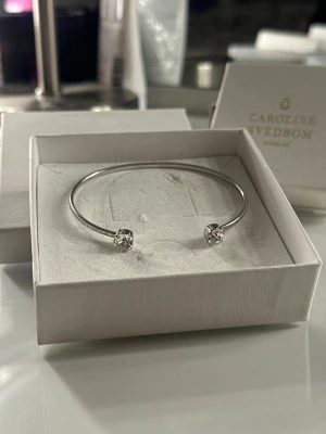 Silverfärgat armband Caroline Svedbom - Stilrent armband i silverfärg från Caroline Svedbom med två gnistrande klara stenar i varje ände. Armbandet är öppet och har en minimalistisk design som passar perfekt till din vardagsstil. Materialet är metall med kristallstenar. Säljer pågrund av att ja bytt från silver till guld! 