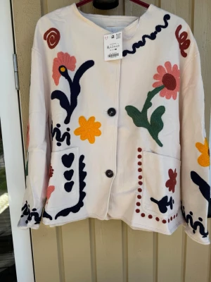 Vit kofta med broderade blommor - Unik vit kofta från Zara med stora broderade blommor i rosa, gult, grönt och marinblått. Koftan har två knappar framtill, stora fickor och ett färgglatt blommotiv även på ryggen. Perfekt för dig som gillar färg och lekfull stil.