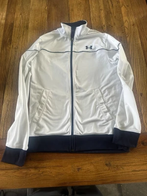 Vit träningsjacka från Under Armour - Snygg vit träningsjacka från Under Armour med mörkblåa detaljer och dragkedja. Jackan har ribbade muddar, två fickor framtill och broderad logga på bröstet. Perfekt för träning eller sportiga outfits.