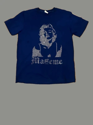 Blå t-shirt med tryck från Masteme - Blå t-shirt från Masteme med ett stort silverfärgat tryck framtill som föreställer ett ansikte och texten 'Masteme'. Klassisk passform och rund halsringning. Perfekt för dig som vill sticka ut med en unik design.