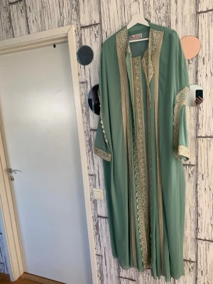 Grön kaftan med guldbroderi - Snygg grön kaftan med detaljerade guldbroderier och pärlbroderier längs kanter och ärmar. Lång och luftig modell med vida ärmar och elegant mönster. Perfekt för dig som vill sticka ut med en unik och traditionell look.