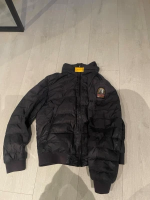 Svart pufferjacka från Parajumpers - Snygg svart pufferjacka från Parajumpers med emblem på ärmen och gul detalj i nacken. Jackan har dragkedja framtill, två sidofickor med dragkedja och ribbade muddar vid ärmslut och nederkant. Perfekt för kalla vinterdagar.