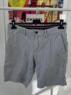 Randiga shorts från Tommy Hilfiger - Snygga randiga shorts från Tommy Hilfiger i vitt och blått. Klassisk design med bälteshällor, knapp och dragkedja framtill. Tillverkade i ett lätt bomullsmaterial, perfekta för varma dagar. Stilsäkra och enkla att matcha med olika outfits. Knappen på ena bakfickan har lossnat men det sitter på extraknappar på insidan så enkelt att åtgärda. 