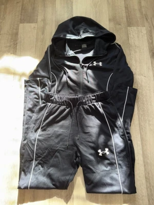 Svart tracksuit från Under Armour - Snyggt svart tracksuit från Under Armour med vit piping. Setet består av en hoodie med dragkedja och matchande byxor med resår i midjan. Båda plaggen har Under Armour-loggan framtill och är gjorda i ett mjukt, sportigt material. Bpda ingår