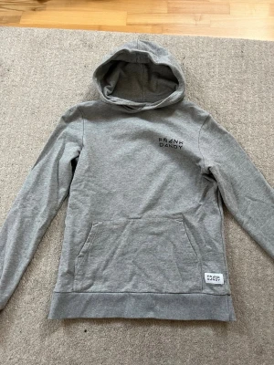 Grå hoodie från Frank Dandy - Snygg grå hoodie från Frank Dandy med svart logga på bröstet och en patch nertill. Klassisk. Perfekt för en avslappnad och stilren känsla. Passform är rätt så small och lite längre, storlek = 14 Y