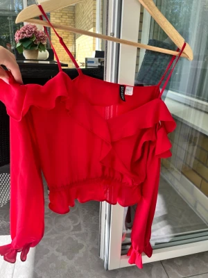 Röd volangblus med omlott - Trendig röd blus från Chiquelle med volangdetaljer och omlott-design. Blusen har långa ärmar med resår vid ärmslut, smala axelband och croppad passform. Perfekt för dig som vill sticka ut med en färgstark topp.