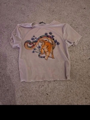 Beige t-shirt med tigertryck - Trendig beige t-shirt med coolt tryck av en tiger och blommor på framsidan. T-shirten har korta ärmar och en rak passform. Perfekt för dig som vill sticka ut med ett unikt motiv. Materialet känns mjukt och lätt mot huden.