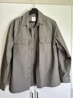 Grå overshirt från H&M - Stilren grå overshirt från H&M med klassisk krage och två bröstfickor med knappar. Rak passform och långa ärmar med knapp vid ärmslut. Perfekt att bära öppen eller stängd över en t-shirt. Gjord i ett mjukt bomullstyg.