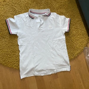 Vit pikétröja från Moncler med röd och blå kant  - Vit pikétröja från Moncler med klassisk krage och knäppning framtill. Röd och blå rand på krage och ärmslut ger en sportig touch. Broderad Moncler-logga på bröstet. Perfekt för en stilren och avslappnad look.