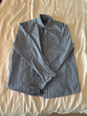 Blårutig skjorta från Hampton Republic - Snygg blårutig skjorta från Hampton Republic med klassisk krage och knäppning framtill. Skjortan har lång ärm, bröstficka och regular fit. Perfekt för en stilren look och tillverkad i mjuk bomull.