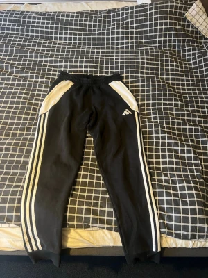 Svarta Adidas mjukisbyxor med vita ränder i storlek S men passar även M - Säljer ett par svarta mjukisbyxor från Adidas med klassiska vita ränder längs benen och vita detaljer vid fickorna. Byxorna har resår i midjan och är tillverkade i mjukt material, perfekta för chill eller träning. Adidas-logga på vänster lår. Väldigt sköna gymbyxor men passar lika bra till fotboll. 