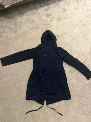Mörkblå parkas från Tommy Hilfiger - Snygg mörkblå parkas från Tommy Hilfiger med huva och dragsko i midjan. Jackan har långa ärmar, två stora fickor framtill och meshfoder på insidan. Perfekt för kyliga dagar och enkel att matcha med olika outfits.