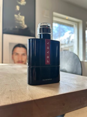 Prada Eau de Parfum Luna rossa 100 ml - Snygg parfym från Prada i en stilren, svart flaska med röd detalj och tydlig logga. Flaskan har en modern, kantig form och genomskinlig kork. Perfekt för dig som gillar exklusiva dofter och vill ha en elegant parfymflaska i badrumshyllan.