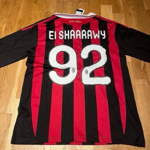 Milan 12/13 long sleeve El Shaarawy #92 - Säljer en långärmad AC Milan matchtröja från Adidas med röda och svarta ränder, vit krage och klubbmärke på bröstet. Tryck med El Shaarawy och nummer 92 på ryggen. Tillverkad i lätt och ventilerande ClimaCool-material.
