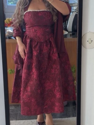 Vinröd långklänning med blommönster - Elegant vinröd långklänning med blommigt mönster och axelbandslös design. Klänningen har en markerad midja och voluminös kjol, samt fina detaljer upptill. Använd endast en gång. 