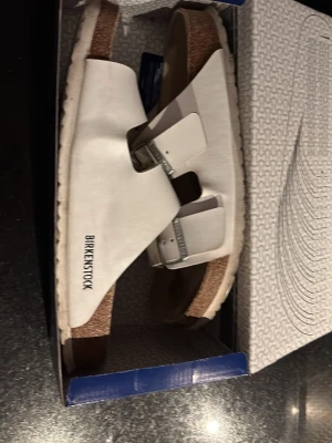 Vita Birkenstock sandaler i läder - Stilrena vita sandaler från Birkenstock med två breda remmar i läder och justerbara metallspännen. Fotbädden är i kork och sulan är vit. Perfekta för en avslappnad och trendig look.
