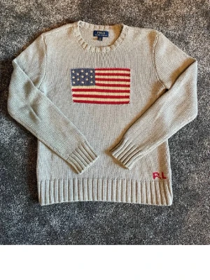 Grå stickad tröja Polo Ralph Lauren - Grå stickad tröja från Polo Ralph Lauren med amerikansk flagga på bröstet och RL-broderi nertill. Tröjan har rund halsringning, långa ärmar och ribbstickade muddar. Perfekt för dig som gillar klassisk och stilren design.
