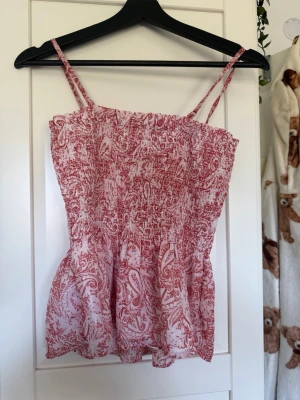 Rosa och vit smockad linnetopp - Supersöt linnetopp med smockad överdel och tunna axelband. Toppen är vit med rosa paisleymönster och har en luftig, lätt form. Perfekt för varma dagar och enkel att styla med jeans eller kjol.