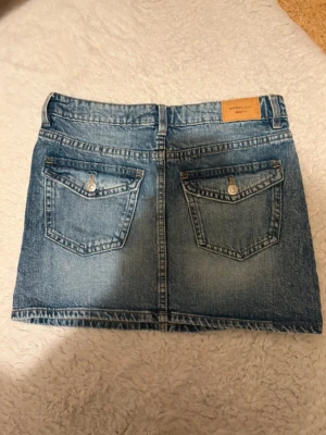 Blå jeanskjol med fickor från Gina tricot  - Säljer en klassisk blå jeanskjol från Gina tricot  Perfect Jeans. Kjolen är kort, har två stora bakfickor med knapp och bälteshällor i midjan. Perfekt till vardags och enkel att matcha med olika toppar. Gjord i slitstarkt denimtyg.