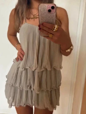 Beige plisserad kortklänning - Superfin beige kortklänning med plisserat tyg och volanger i flera lager. Klänningen är ärmlös och har en luftig, flowy passform som ger en riktigt snygg och avslappnad look. Perfekt för varma dagar och somriga vibes.