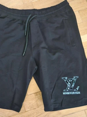 Svarta Louis Vuitton shorts med snörning - Svarta shorts från Louis Vuitton med elastisk midja och snörning. Stort LV-logotyptryck i vitt och blått på ena benet. Perfekta för en avslappnad och trendig stil.