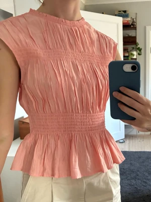 Rosa plisserad topp med volang - Söt ljusrosa lite korallig topp med plisserat tyg och volang nertill. Modellen är ärmlös med rund halsringning och smockad midja som ger en snygg siluett. Så söt verkligen!