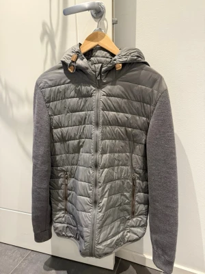 Grå cardigan med stickade ärmar - Snygg grå pufferjacka med quiltat mönster och huva. Ärmarna är stickade i en matchande grå nyans och jackan har dragkedja framtill samt två fickor med dragkedja. Perfekt för kalla dagar och har coola detaljer vid huvan.
