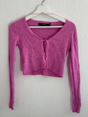Ribbad rosa croppad topp Vero Moda - Trendig ribbad topp från Vero Moda i en härlig rosa färg. Modellen är croppad med lång ärm, v-ringning och knyt framtill. Perfekt för att lyfta din outfit med en poppig färg och snygg passform.