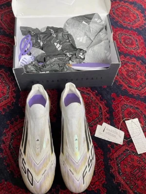 Adidas F50 fotbollsskor vita/lila - Säljer ett par Adidas F50 fotbollsskor i vitt med lila detaljer och svarta ränder. Skorna har en strömlinjeformad design utan snörning och är tillverkade i lätt syntetmaterial för maximal fart på planen. Originalkartong och extra skosnören medföljer.