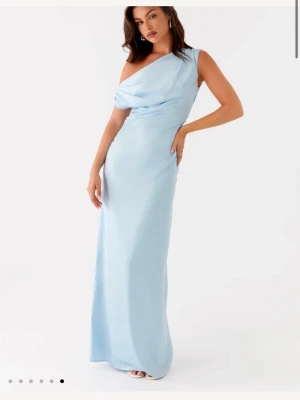 Ljusblå långklänning one shoulder - Elegant ljusblå långklänning med one shoulder-design och draperad överdel. Klänningen har en smickrande, figurnära passform och är tillverkad i ett glansigt, mjukt material som ger en lyxig känsla. Perfekt för speciella tillfällen när du vill sticka ut.