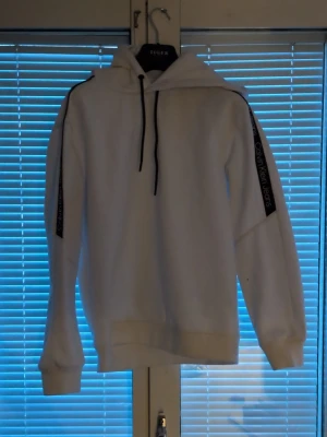 Vit hoodie från Calvin Klein Jeans - Snygg vit hoodie från Calvin Klein Jeans med svarta detaljer och logga längs ärmarna. Klassisk huva med svarta dragsnören och ribbade muddar. Perfekt för en clean och stilren look.
