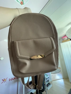 Beige ryggsäck från Primark - Snygg beige ryggsäck från Primark i fejkläder med guldfärgad detalj på framfickan. Ryggsäcken har ett stort huvudfack med dragkedja och en mindre innerficka. Perfekt för dig som vill ha en stilren och enkel väska till vardagen.