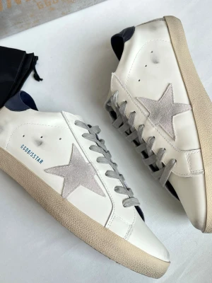 GGDB Superstar vita sneakers - Snygga GGDB Superstar sneakers i vitt läder med grå stjärndetalj på sidan och mörkblå häl. Grå skosnören och beige sula ger en cool kontrast. Klassisk låg modell med tydlig branding och retrovibe. Perfekta för dig som gillar streetstyle och unika detaljer. Dustbag medföljer, skriv vid funderingar!