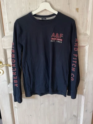 Mörkblå long sleeve från Abercrombie & Fitch - Mörkblå långärmad t-shirt från Abercrombie & Fitch med tryck i rött och vitt på bröstet och längs båda ärmarna. Klassisk rund halsringning och skön passform. Perfekt för en avslappnad och stilren look.