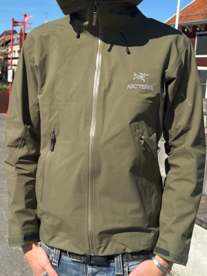 Olivgrön vindjacka från Arc'teryx - Snygg olivgrön vindjacka från Arc'teryx med huva och hel dragkedja framtill. Jackan har två dragkedjeförsedda sidofickor och logga på bröstet. Perfekt för blåsigt väder, regn eller bara allmänt en skön jacka som funkar i många sammanhang🌊☀️