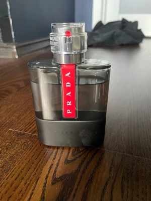 Prada Luna Rossa Carbon parfym - Snygg parfymflaska från Prada Luna Rossa Carbon med modern design. Flaskan är svart och grå med en röd detalj där märket står. Tillverkad i glas och plast, passar perfekt för dig som gillar exklusiva dofter.