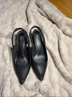 Svarta pumps XIT - Säljer ett par svarta pumps från XIT med spetsig tå och bred blockklack. Skorna har öppen häl med elastiskt band för skön passform. Tillverkade i ett slätt material som ger en stilren look. Perfekta för dig som gillar klassiska men trendiga skor. Lite slitage vilket visas.