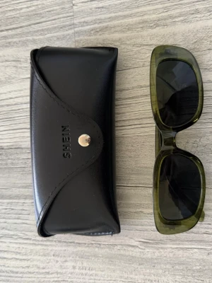 Gröna solglasögon från SHEIN - Snygga solglasögon från SHEIN med rektangulära bågar i transparent grön plast. Kommer med ett svart fodral i syntetmaterial med tryckknapp. Perfekt accessoar för att lyfta din stil i solen.