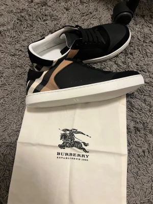 Burrbery skor - Snygga låga sneakers från Burberry i svart med klassiskt beige, vitt och svart rutmönster på sidan. Skorna har vita sulor, svarta skosnören och detaljer i både textil och läder. Perfekta för dig som vill ha en stilren och lyxig look.