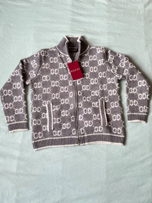 Grå ziptröja med GG-mönster från Gucci - Snygg grå ziptröja från Gucci med vitt GG-monogram över hela tröjan. Tröjan har hög krage, ribbade muddar och fickor framtill. Tillverkad i mjukt stickat material och har vita detaljer längs kanterna. Perfekt för dig som gillar exklusiv streetstyle.