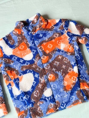 Färgglad teddyjacka med mönster - Mysig teddyjacka i blått, orange, brunt och vitt med abstrakt mönster. Jackan har hög krage och dragkedja framtill. Perfekt för dig som vill sticka ut och hålla dig varm under kyliga dagar. Materialet är fluffigt och mjukt för maximal komfort.