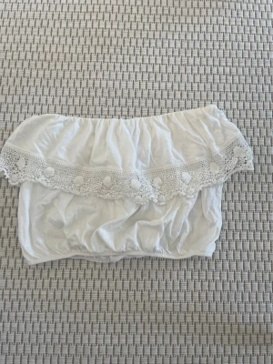 Vit offshoulder topp från New Look - Söt vit offshoulder topp från New Look med virkade spetsdetaljer och små bollar längs kanten. Toppen är croppad och har resår både upptill och nedtill för en bekväm passform. Perfekt för sommarens alla tillfällen!