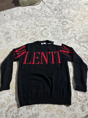 Svart tröja VALENTINO  - Svart stickad tröja från Valentino med stor röd logotyp som sträcker sig över bröstet och ut på ärmarna. Tröjan har rund halsringning och ribbade muddar vid ärmslut och nederkant. Perfekt för dig som gillar statement-plagg.