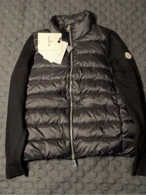 Svart dunväst från Moncler - Snygg svart dunväst från Moncler med quiltad framsida och hög krage. Västen har dragkedja framtill och Moncler-logga på ärmen. Materialet är glansigt och fyllt med dun, vilket ger en lyxig känsla. Perfekt för lager-på-lager och streetstyle.
