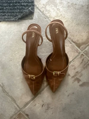 Bruna spetsiga pumps från Zara - Snygga bruna pumps från Zara med spetsig tå, smal klack och dekorativt spänne i guld framtill.  Använda 1 gång 