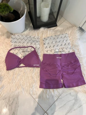 Slutsåld färg från DFYNE - Säljer ett snyggt lila träningsset från DFYNE som består av en sport-bh med twistad front och smala axelband samt matchande shorts med hög midja. Materialet är stretchigt och bekvämt, perfekt för gymmet eller yoga. Logotypen finns på både bh och shorts.impact 4.5